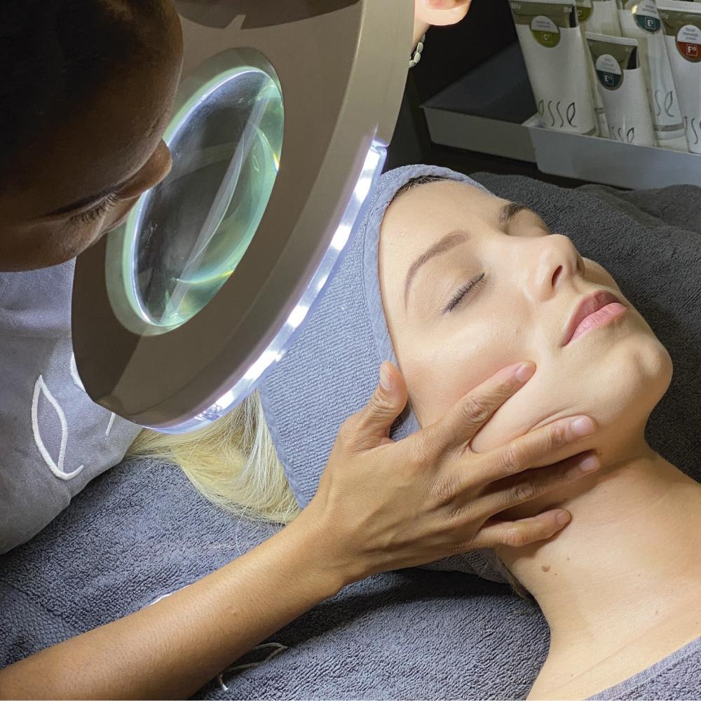 A person is receiving a facial treatment at a spa. Another individual is gently holding their face and examining it under a large magnifying lamp with built-in lights. Skincare products are visible in the background.