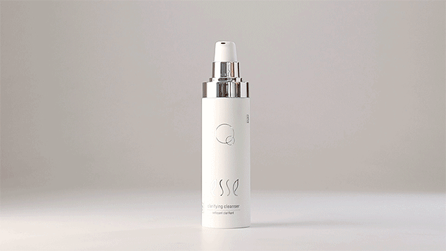 A white pump bottle with silver accents, labeled "Esse Clarifying Cleanser," stands on a light gray surface. The bottle features a logo and text detailing, with a minimalist design against a neutral background.
