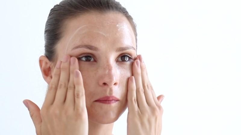 With their hair pulled back, a person gently massages their face with both hands while applying Esse Gel Cleanser. The background is a soft white, and they appear focused on their skincare routine.