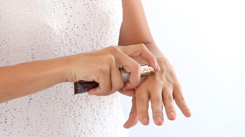A person wearing a white lace dress applies Esse Hand Cream from a tube onto the back of their hand. The image focuses on their hands and the tube, with a light background.