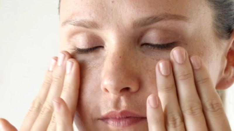 A close-up of a person with closed eyes gently massaging their face with both hands, focusing on the area around their eyes with Esse Intensity Serum. The skin appears clear and smooth, and the person's fingernails are manicured with a light, neutral polish. The background is softly blurred.