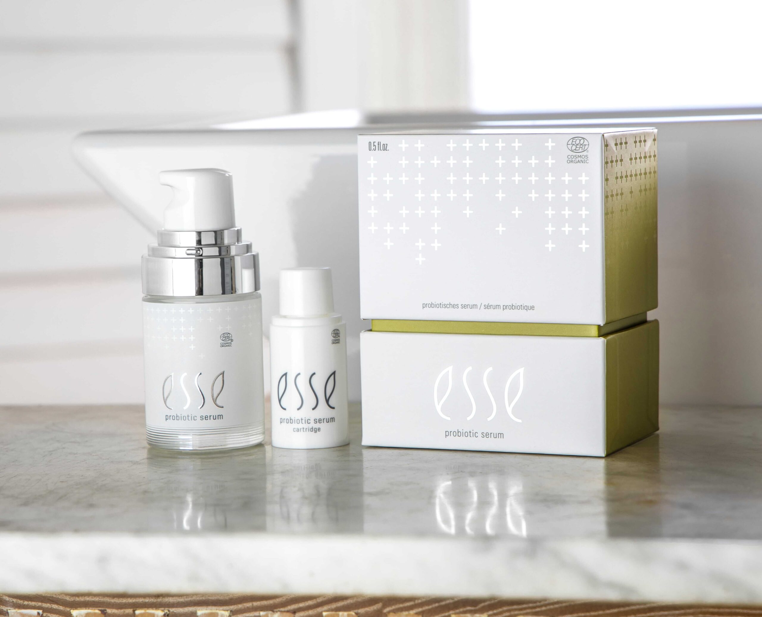 Two bottles of Esse Probiotic Serum sit on a marble surface beside their white and green boxes, with a softly bright, blurred background.