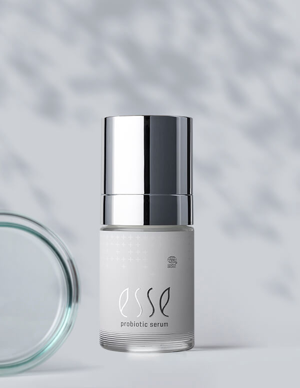 A bottle of Esse Probiotic Serum with a shiny silver cap stands upright on a light surface. The white label is minimalist, and part of a glass lid appears to the left. Soft shadows create patterns in the background.